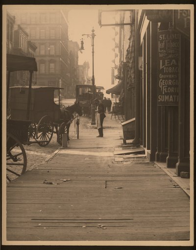 Vertikales Schwarz-Weiß-Foto von pferdegezogenen Fahrzeugen, die an einem Holzplanken-Bürgersteig parken. Ladenfronten säumen den Bürgersteig und ein Mann steht an der Ecke von William G. Swekosky